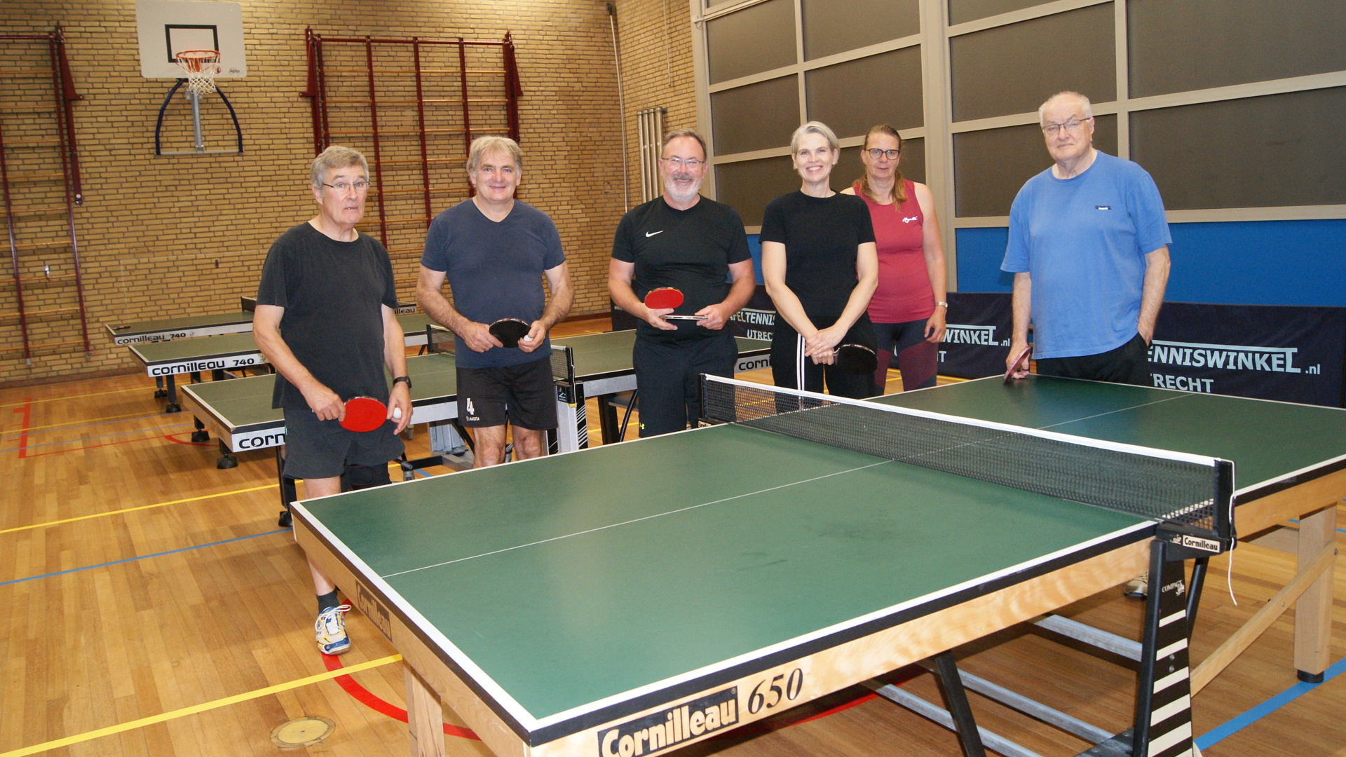 Groep met mensen met Parkinson poseert tijdens de les van tafeltennisvereniging Pegasus bij tafeltennistafel.
